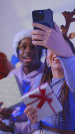 Foto-De-Estudio-De-Vídeo-Vertical-De-Amigos-De-La-Generación-Z-En-Navidad-Sentados-En-Un-Sofá-Con-Gorro-De-Papá-Noel-Y-Astas-De-Reno-Tomándose-Un-Selfie-En-Un-Teléfono-Móvil